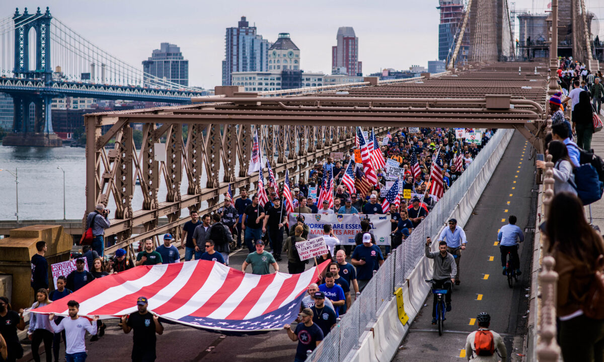 Funcionários municipais seguram cartazes e afirmam slogans enquanto marcham pela ponte do Brooklyn durante um protesto contra o mandato de vacinação para a COVID-19, em Nova Iorque, no dia 25 de outubro de 2021 (Ed Jones/AFP via Getty Images)