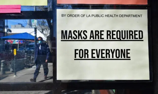 Um homem usando uma máscara facial protetora passa por uma placa exigindo máscaras postadas em uma loja em Los Angeles, Califórnia, em 2 de março de 2022. (Frederic J. Brown/AFP via Getty Images)
